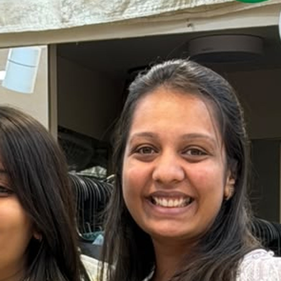 A close-up selfie-style photo of a smiling woman with dark hair, with another person partially visible at the left edge and an indoor/outdoor doorway background.
