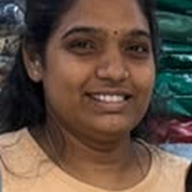A smiling woman wearing a beige and brown color-blocked t-shirt, standing next to a young girl in front of a background of stacked green and red bags.