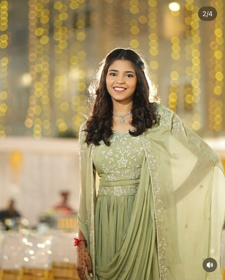A woman wearing a light sage green embroidered traditional outfit with a matching sheer shawl, standing in front of a warm bokeh background. The photo appears to be from an Instagram-style feed with visible interface elements and user tags.
