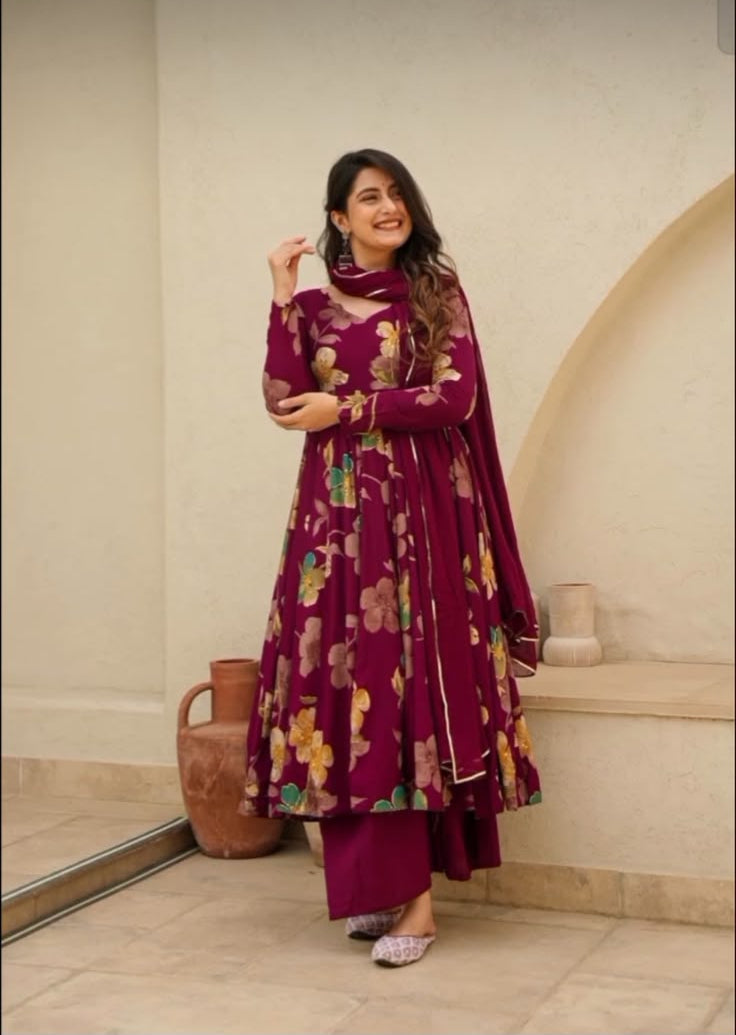 A woman modeling a deep magenta floral printed traditional outfit with a matching dupatta in an indoor setting with beige walls and arches.