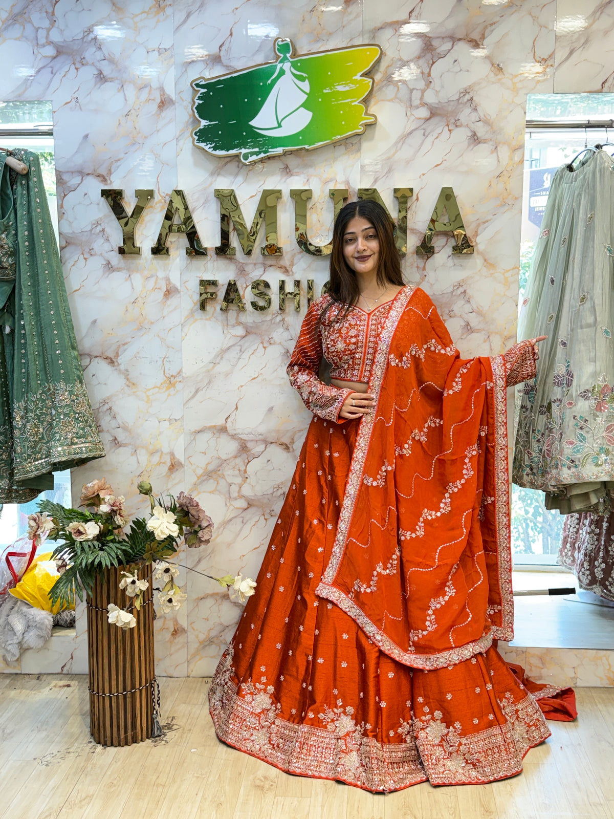 A woman models a vibrant orange-red embroidered traditional Indian outfit with a matching dupatta in a boutique setting. The background features a marble wall with the brand name 'YAMUNA FASHION'.